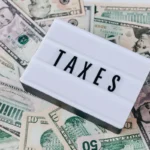 Tulisan TAXES di atas tumpukan uang dolar, menggambarkan konsep fiskal dan kebijakan penerimaan negara dari pajak